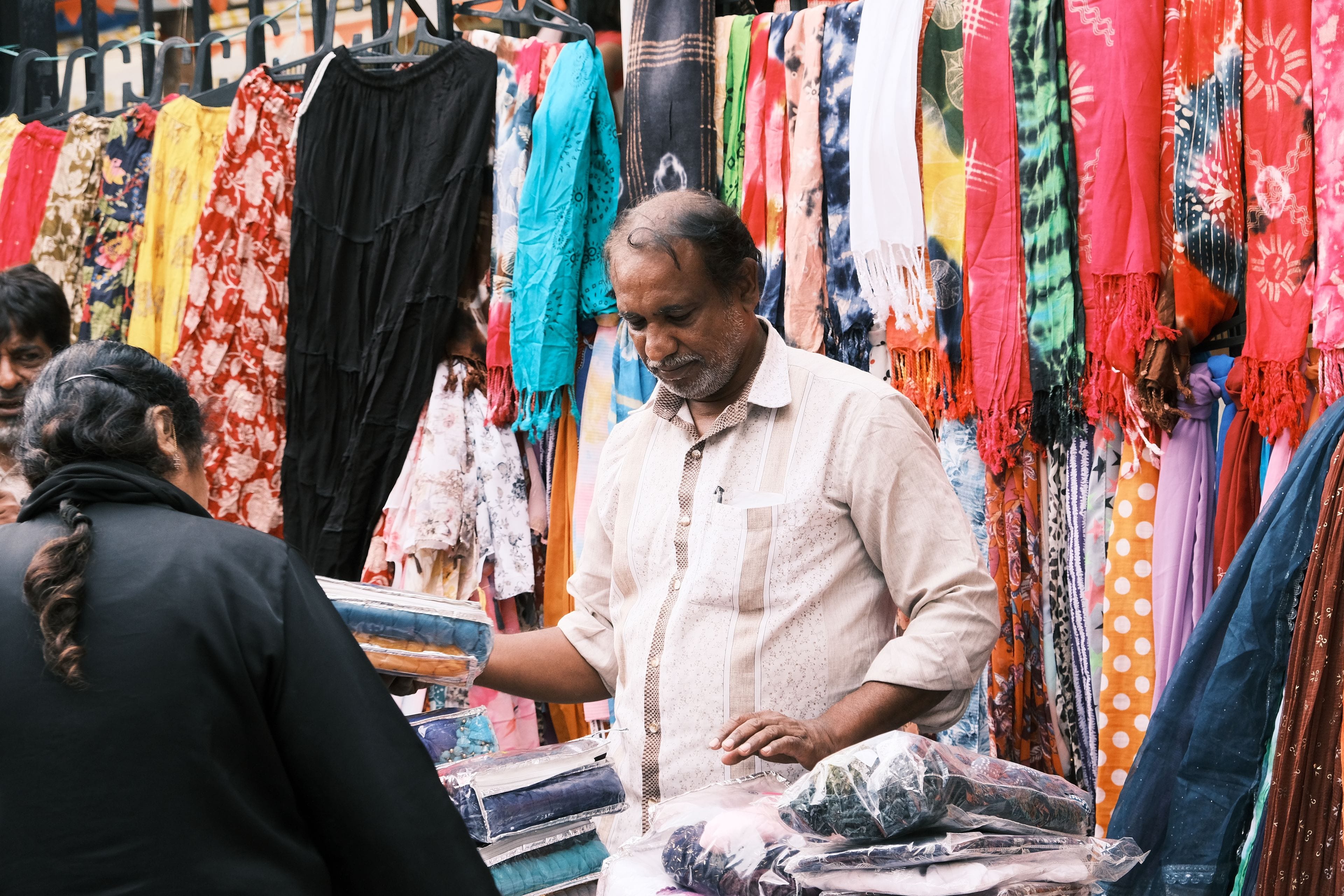 A shopkeeper serving a customer across a counter stacked with folded clothes